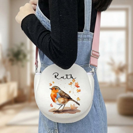 A person wearing denim overalls and a black long-sleeve top carries a round white crossbody bag with a pink strap. The bag features a printed illustration of a robin bird with the name 'Ruth' above it and small autumn leaves around it.