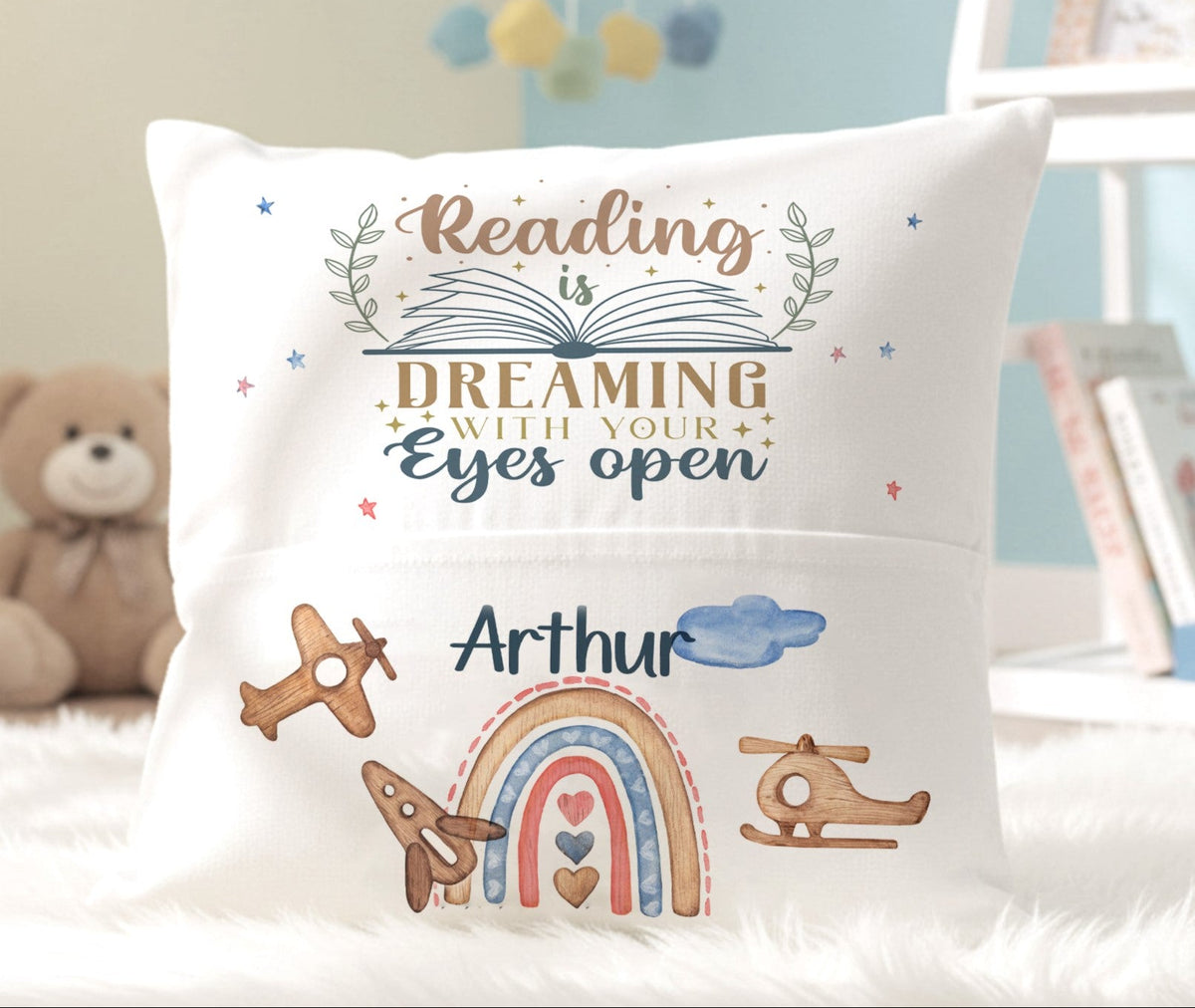 A square white throw pillow with a custom printed design that reads 'Reading is DREAMING with your Eyes open' and is personalized with the name 'Arthur' above a watercolor rainbow, airplanes, and helicopters. The pillow is styled on a white faux fur surface with a teddy bear and books in the background.