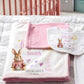 Personalized baby blanket and bib set with a bunny illustration and the name Smith, laid out on a white quilted bedspread.