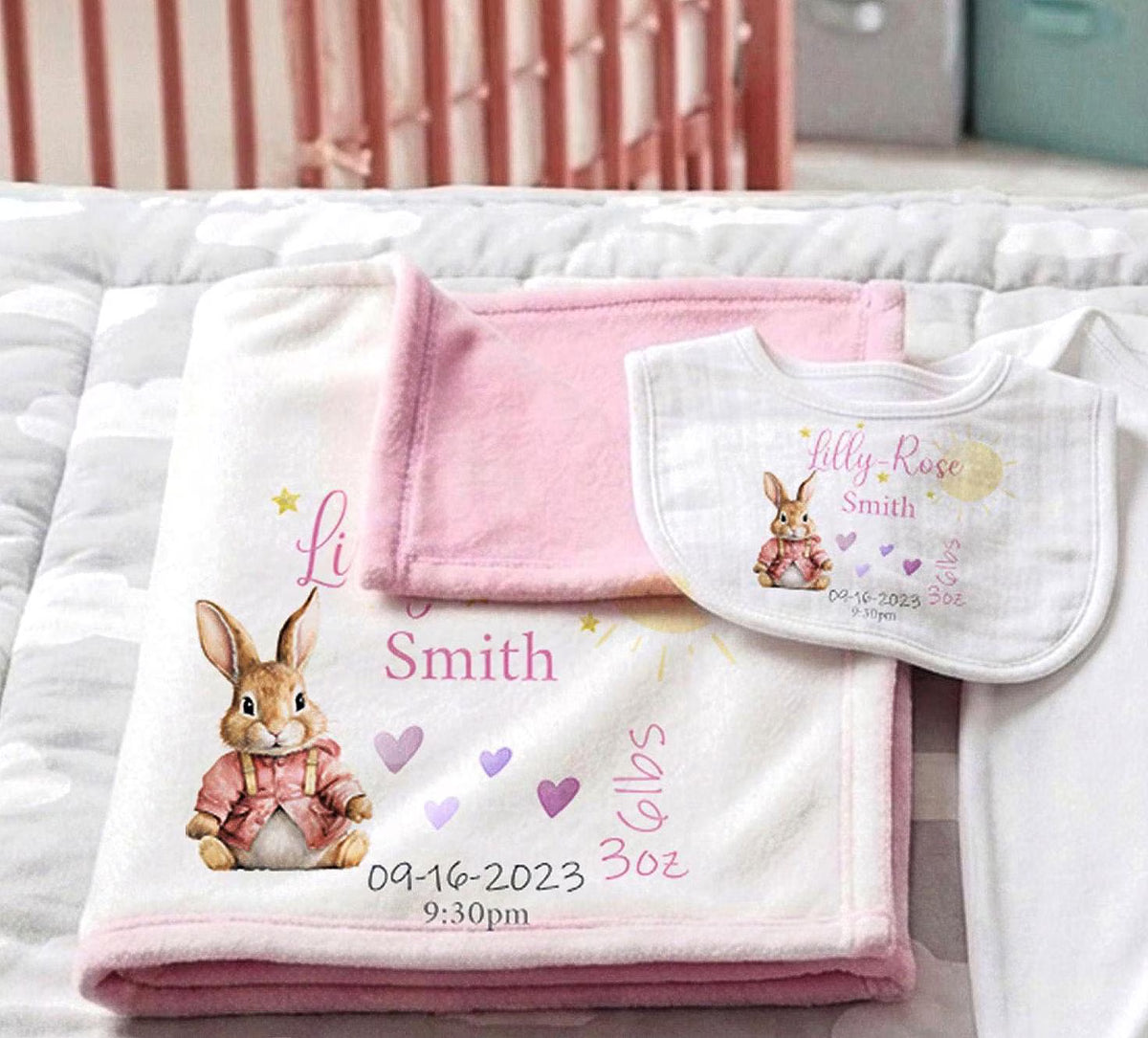 Personalized baby blanket and bib set with a bunny illustration and the name Smith, laid out on a white quilted bedspread.
