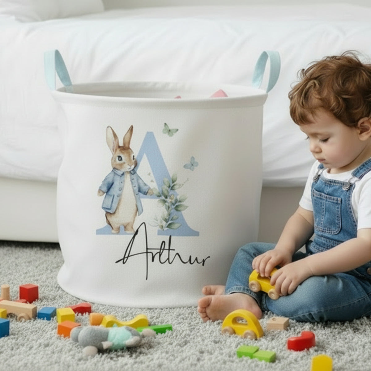A child in denim overalls sits on a gray carpet playing with toys beside a large white fabric storage bin printed with a bunny, a blue letter A, butterflies, and the name Arthur. The room has white bedding in the background.