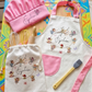 A children's cooking-themed play set arranged on a colorful table, including a pink chef hat, white apron with pink trim and embroidered characters, a matching drawstring bag, wooden spoons, a whisk, and a silicone brush. The set is photographed in a bright room with toys and shelves in the background.