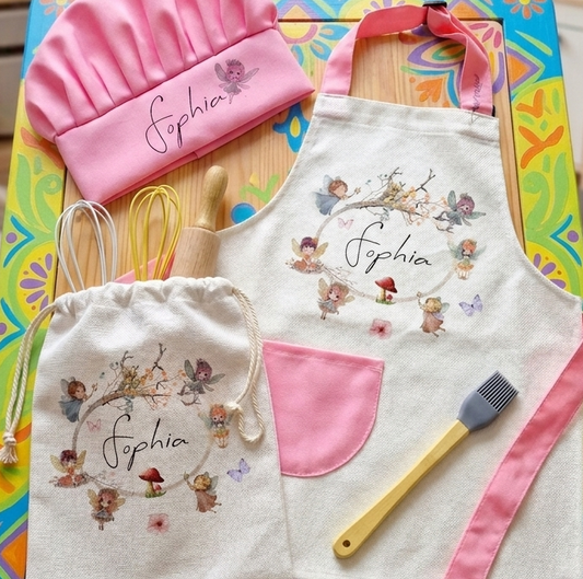 A children's cooking-themed play set arranged on a colorful table, including a pink chef hat, white apron with pink trim and embroidered characters, a matching drawstring bag, wooden spoons, a whisk, and a silicone brush. The set is photographed in a bright room with toys and shelves in the background.