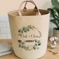 A beige fabric laundry hamper with two handles sits on a wooden table in a styled room. The front of the hamper has a personalized floral wreath design with the text 'Jack & Olivia's LAUNDRY'.