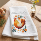 White towel with rooster design and 'Julia's Kitchen' text on a wooden table with eggs and flowers.