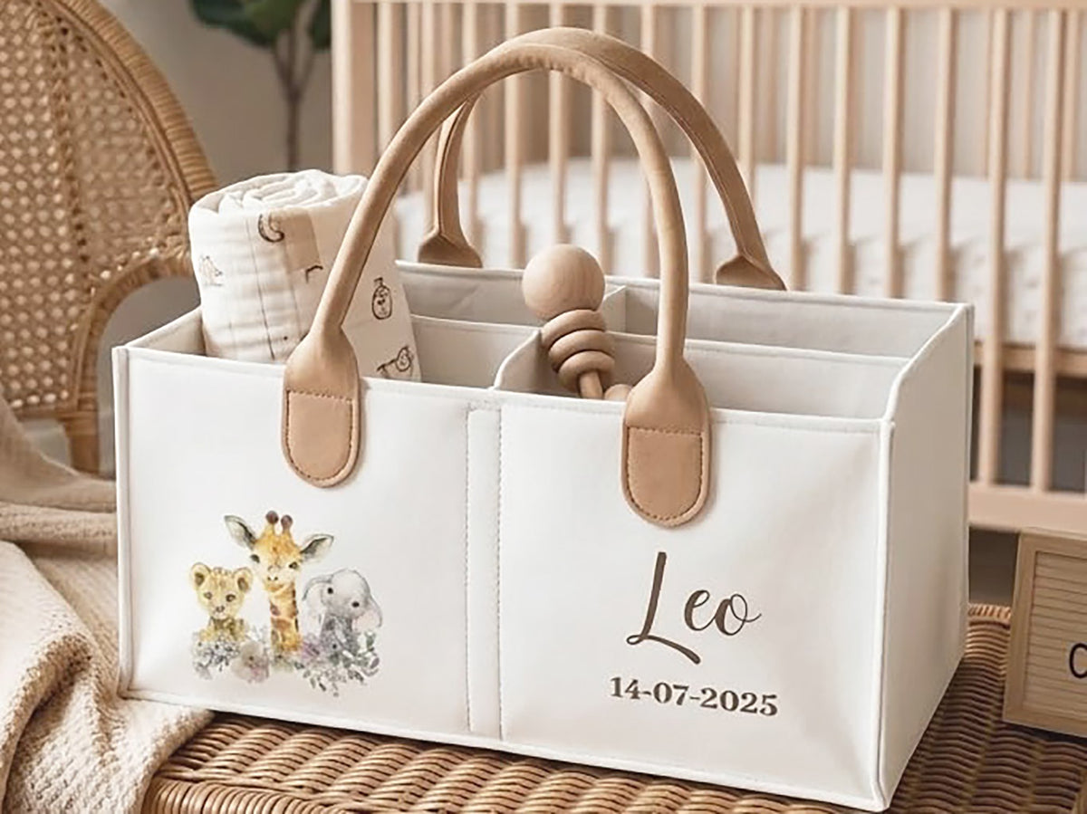 A cream fabric baby caddy organizer with tan handles, shown in a nursery setting. The organizer features a watercolor print of a giraffe, lion, and elephant on the left, and a personalized name 'Leo' with the date '14-07-2025' printed on the right.