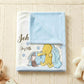 A folded baby blanket with a light blue reverse side and a printed cartoon illustration of Winnie the Pooh hugging a blue bunny, placed on a cream cable-knit blanket with a plush teddy bear and a small stuffed rabbit prop.