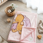 A folded pink plush blanket featuring Winnie the Pooh and a purple pillow graphic, styled on a light wood table with a small wicker basket, cotton boll, wooden blocks, and a drawstring pouch.