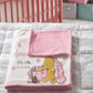 A personalized baby blanket with a pink and white cloud pattern and a cartoon character print, folded on a gray cloud-patterned quilt in a nursery setting with a pink crib in the background.