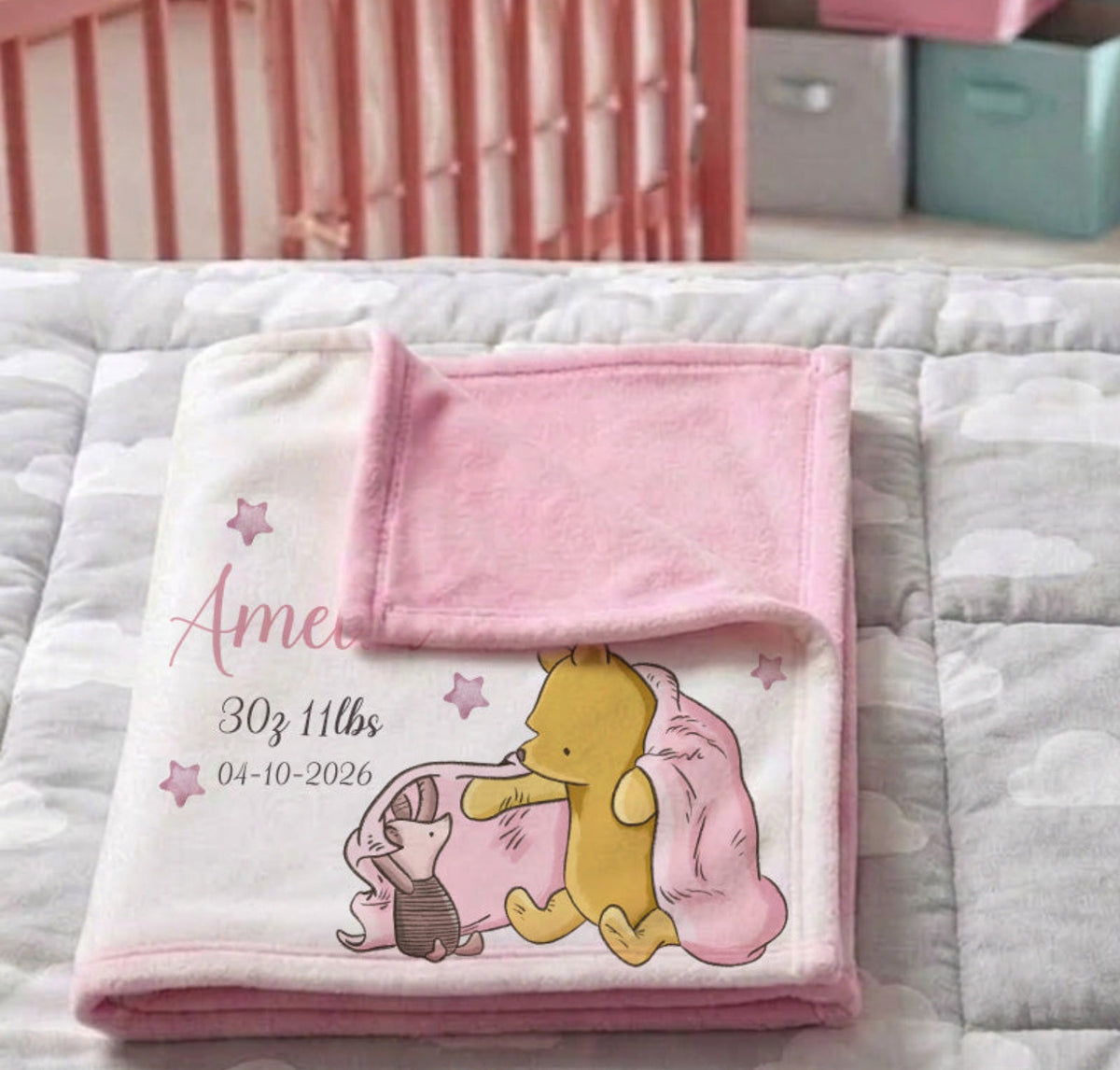 A personalized baby blanket with a pink and white cloud pattern and a cartoon character print, folded on a gray cloud-patterned quilt in a nursery setting with a pink crib in the background.
