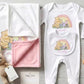 A baby gift set laid out on a light wood surface, including a white long-sleeve footed bodysuit with a Winnie the Pooh and Piglet rainbow graphic and the name 'Emily' printed on the chest pocket, a white and pink blanket with a Winnie the Pooh print, a small woven bunny toy, and a wooden baby rattle.