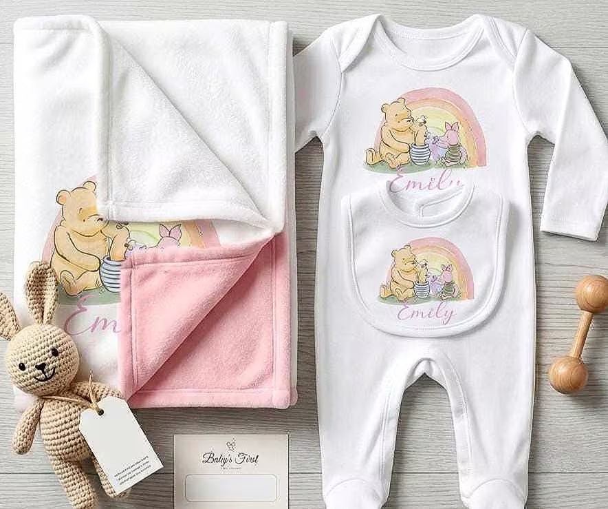 A baby gift set laid out on a light wood surface, including a white long-sleeve footed bodysuit with a Winnie the Pooh and Piglet rainbow graphic and the name 'Emily' printed on the chest pocket, a white and pink blanket with a Winnie the Pooh print, a small woven bunny toy, and a wooden baby rattle.