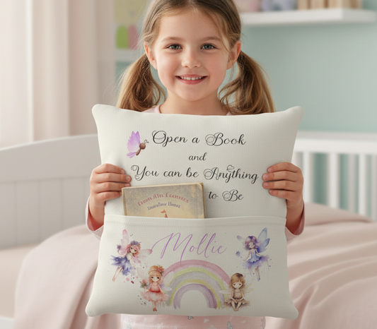 A young girl with pigtails smiles while holding a personalized white pillow in a bright, pastel-colored bedroom. The pillow features the printed phrase 'Open a Book and You Can Be Anything To Be' on the top half and a pocket on the bottom decorated with a purple fairy, a small bunny, and the name 'Mollie' next to a rainbow illustration.
