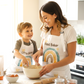 A woman and a young boy baking together in a bright kitchen, both wearing white aprons with a colorful rainbow design and the words 'Head Baker' and 'Little Baker' respectively.