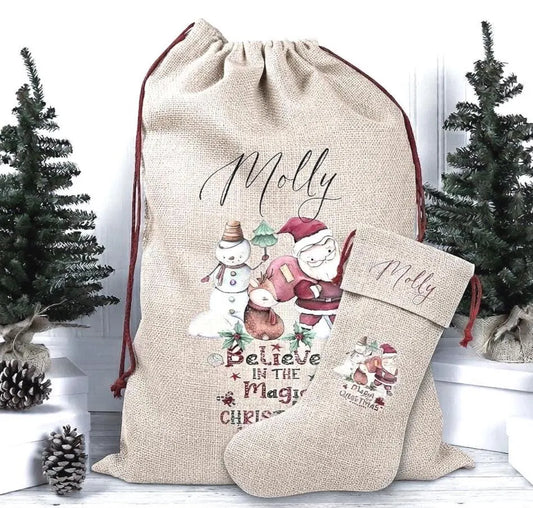 A personalized Christmas gift sack and matching stocking set in natural burlap, printed with the name Molly, a snowman, Santa, and the phrase 'Believe in the Magic of Christmas.' The set is styled with small artificial Christmas trees and pinecones on a white surface against a white background.