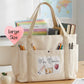 A large beige canvas tote bag with multiple pockets, shown on a wooden desk in a classroom setting. The bag features a front pocket with a 'Mrs.