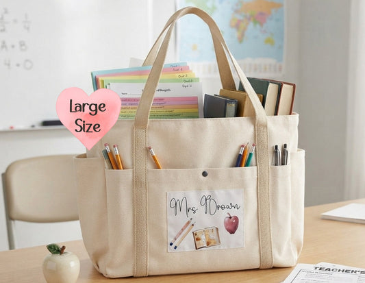 A large beige canvas tote bag with multiple pockets, shown on a wooden desk in a classroom setting. The bag features a front pocket with a 'Mrs.