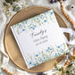A personalized hardcover notebook with a white cover decorated with blue and green watercolor wildflowers, featuring the text 'Emily's Hen Party 20-05-2024' in black script. The notebook is styled in a flat-lay arrangement on a round woven straw placemat, surrounded by wooden beads, white flowers, and green leaves on a draped cream fabric background.