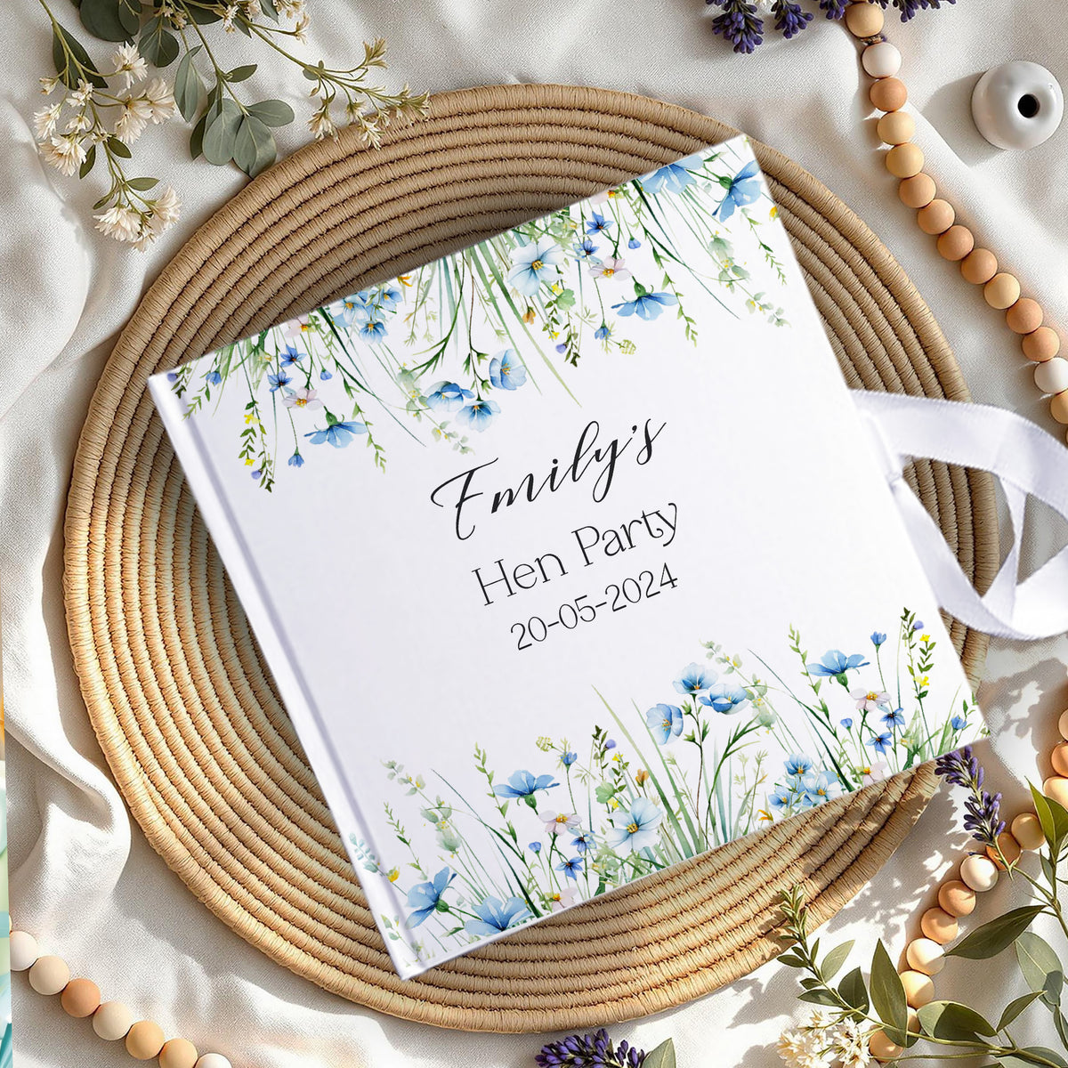 A personalized hardcover notebook with a white cover decorated with blue and green watercolor wildflowers, featuring the text 'Emily's Hen Party 20-05-2024' in black script. The notebook is styled in a flat-lay arrangement on a round woven straw placemat, surrounded by wooden beads, white flowers, and green leaves on a draped cream fabric background.
