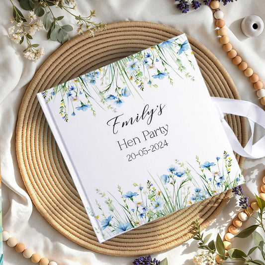 A personalized hardcover notebook with a white cover decorated with blue and green watercolor wildflowers, featuring the text 'Emily's Hen Party 20-05-2024' in black script. The notebook is styled in a flat-lay arrangement on a round woven straw placemat, surrounded by wooden beads, white flowers, and green leaves on a draped cream fabric background.