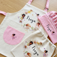 A personalized children's fabric apron set laid flat on a light wood surface, including a cream apron with pink straps and pocket, a matching cream drawstring bag, and a pink drawstring bag. The apron and bag are printed with a fairy-themed circular illustration and the name 'Freya' in black script.