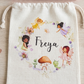 A personalized cream-colored fabric drawstring pouch with a fairy-themed illustration and the name 'Freya' in black script, displayed on a light wood surface with a pink ruffled band above it.