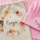 A pink children's apron set laid flat on a round wooden surface, including a pink apron with fairy illustrations and the name 'Freya,' a cream drawstring pouch, and a matching pink chef hat. The set is photographed in a styled flat-lay arrangement with no person or real environment beyond the tabletop backdrop.