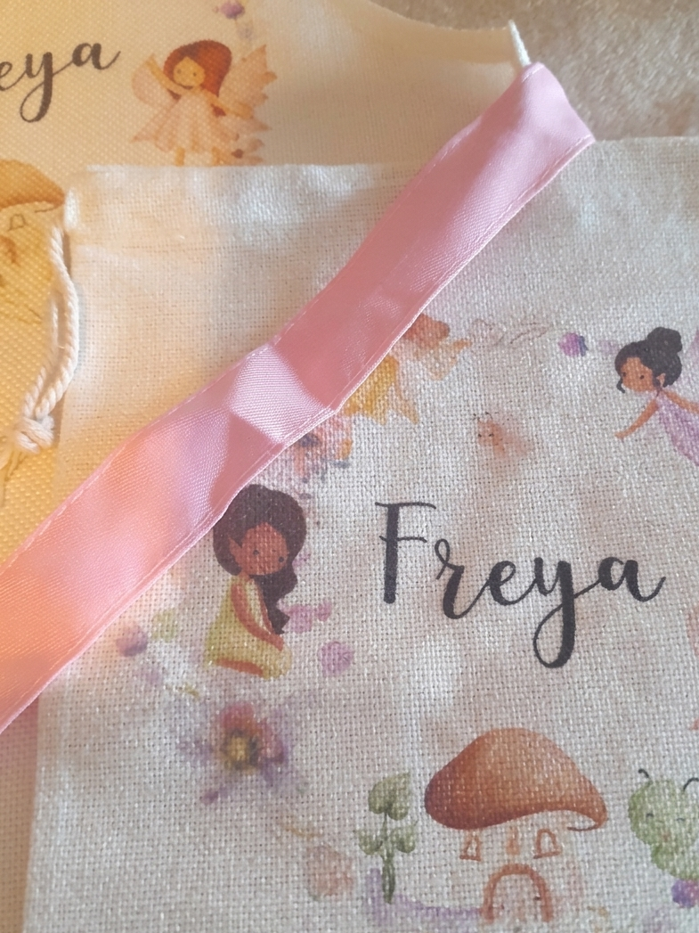 A close-up of a personalized baby blanket featuring the name 'Freya' in black script, surrounded by watercolor illustrations of fairies, a mushroom house, and small flowers. The blanket is decorated with a wide pink satin ribbon and is laid on top of another blanket.