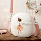 A personalized round canvas shoulder bag in white with a pink strap, featuring an illustration of a ballerina in a pink tutu and the name 'Mallie' printed on the front, displayed on a wooden table in a cozy room setting.