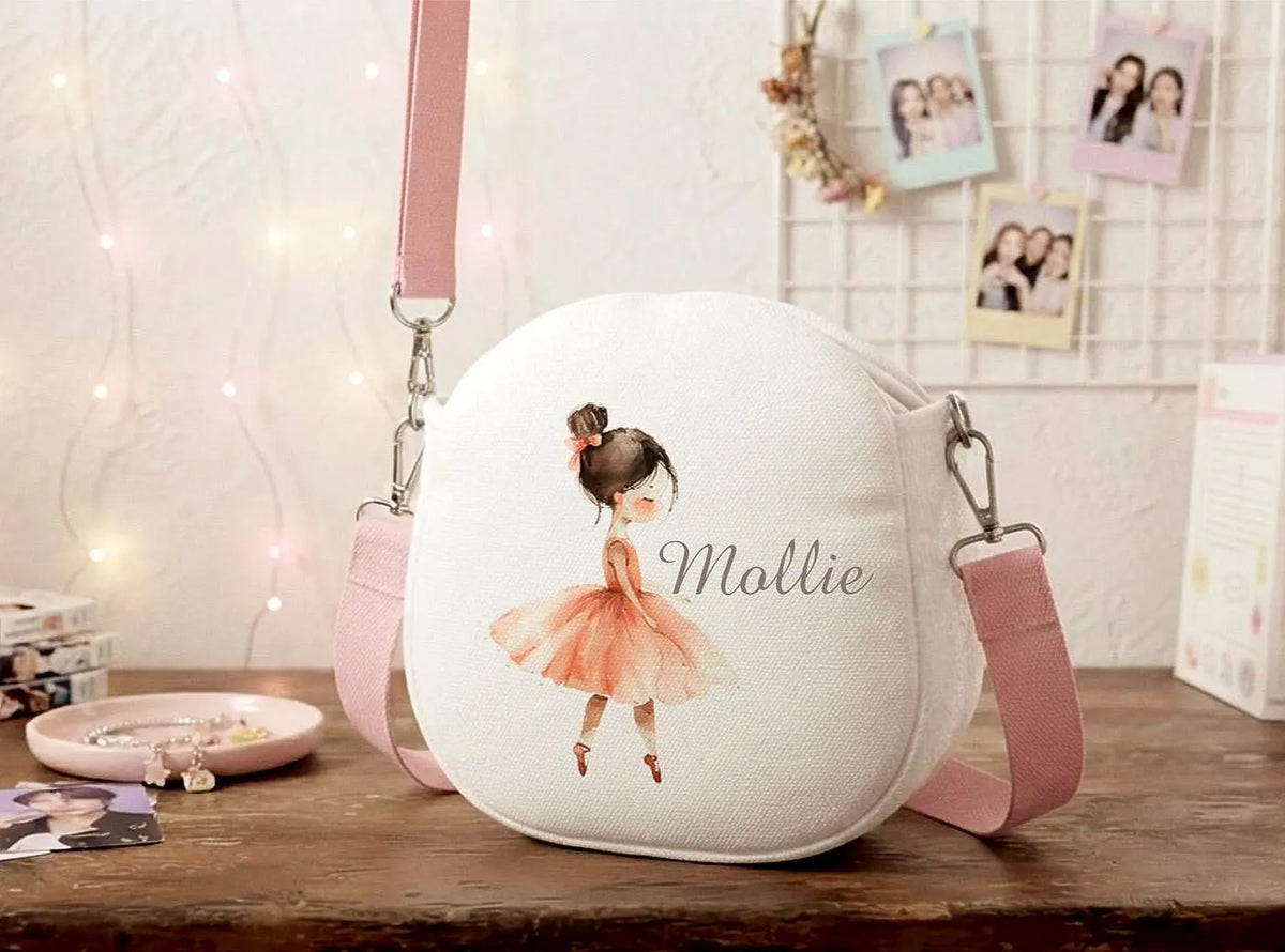 A personalized round canvas shoulder bag in white with a pink strap, featuring an illustration of a ballerina in a pink tutu and the name 'Mallie' printed on the front, displayed on a wooden table in a cozy room setting.
