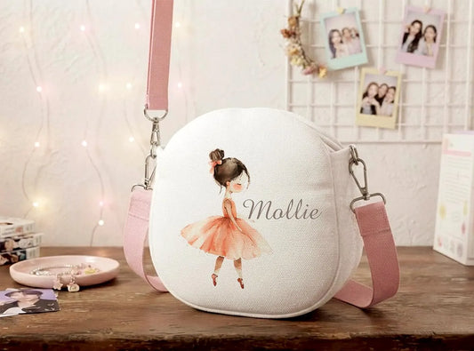 A personalized round canvas shoulder bag in white with a pink strap, featuring an illustration of a ballerina in a pink tutu and the name 'Mallie' printed on the front, displayed on a wooden table in a cozy room setting.
