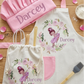 A personalized cooking-themed set laid out on a wooden surface, including a pink chef hat, a cream apron with a floral fairy print and the name 'Darcey', a matching drawstring bag, and two silicone kitchen brushes.