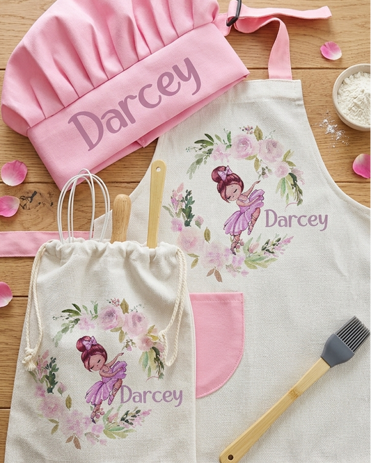A personalized cooking-themed set laid out on a wooden surface, including a pink chef hat, a cream apron with a floral fairy print and the name 'Darcey', a matching drawstring bag, and two silicone kitchen brushes.