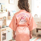 A child viewed from behind wearing a peach satin birthday robe with 'Birthday Girl Emily 2025' printed on the back, standing in a brightly lit children's bedroom.