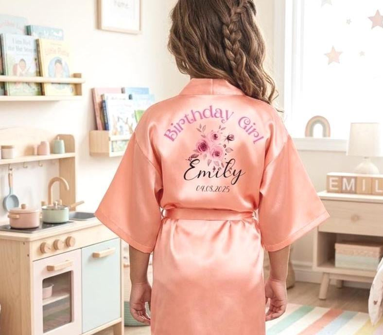 A child viewed from behind wearing a peach satin birthday robe with 'Birthday Girl Emily 2025' printed on the back, standing in a brightly lit children's bedroom.