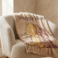 A cozy beige armchair in a warm room setting with sunlight, draped with a plush throw blanket printed with the words 'Snuggle Blank' and a cartoon bear illustration. A small wooden side table with a mug and a potted plant sits to the right.