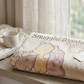 A folded cream-colored plush blanket with a Winnie the Pooh bear design and the words 'Snuggle Blanket' printed on it, resting on a wooden windowsill next to a white mug. A window with sheer curtains and a blurred green garden background are visible.