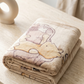 A folded plush blanket with a cartoon bear print in soft beige, tan, and muted pink tones, resting on a light stone surface.