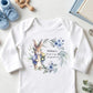A white long-sleeve baby bodysuit laid flat on a light wood surface, featuring a watercolor-style illustration of a rabbit in a floral wreath with the text 'William's 1st Birthday 3rd September 2023.' Blue knit booties, wooden toys, a spoon, a book, and eucalyptus leaves surround the bodysuit as styled props.