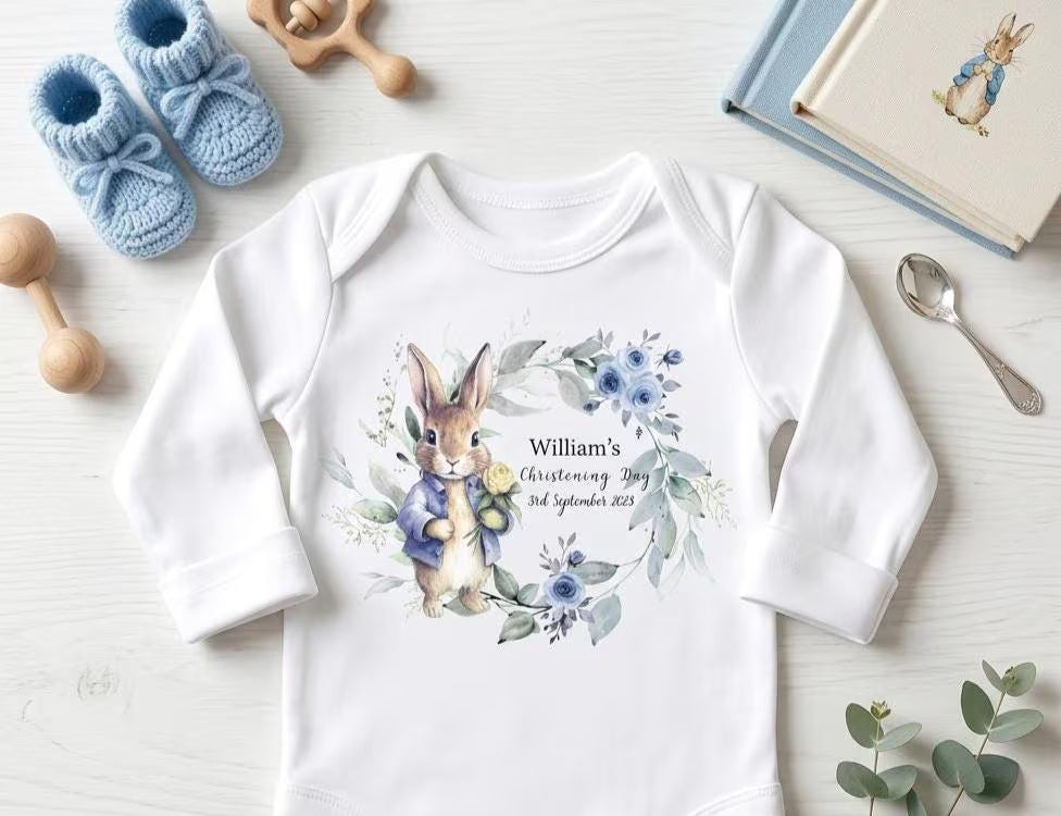 A white long-sleeve baby bodysuit laid flat on a light wood surface, featuring a watercolor-style illustration of a rabbit in a floral wreath with the text 'William's 1st Birthday 3rd September 2023.' Blue knit booties, wooden toys, a spoon, a book, and eucalyptus leaves surround the bodysuit as styled props.