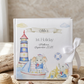 A personalized children's holiday keepsake photo book with a white cover, decorated with watercolor illustrations of a lighthouse, sailboat, elephant, and beach elements. The book is styled on a rustic wooden surface with a white ribbon bow, a small straw hat, seashells, and sandals, set against a warm, neutral background.