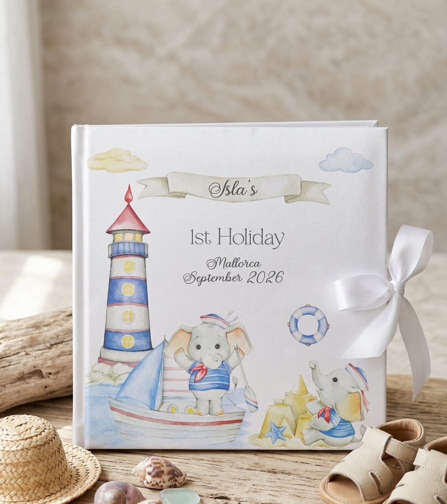 A personalized children's holiday keepsake photo book with a white cover, decorated with watercolor illustrations of a lighthouse, sailboat, elephant, and beach elements. The book is styled on a rustic wooden surface with a white ribbon bow, a small straw hat, seashells, and sandals, set against a warm, neutral background.