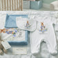A personalized baby bedding and clothing set arranged on a quilted bed in a nursery. The set includes a white footed sleeper with a bunny and floral print and the name 'William', a matching bib, folded blue and white blankets, a stuffed bunny, a teddy bear, and a wooden rattle.