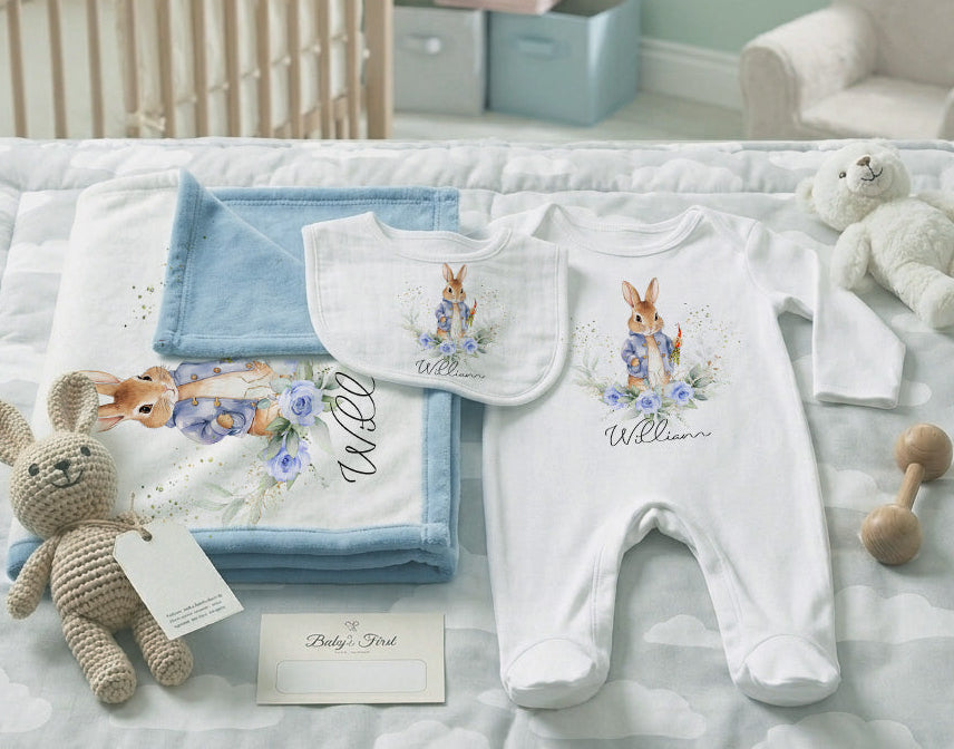 A personalized baby bedding and clothing set arranged on a quilted bed in a nursery. The set includes a white footed sleeper with a bunny and floral print and the name 'William', a matching bib, folded blue and white blankets, a stuffed bunny, a teddy bear, and a wooden rattle.