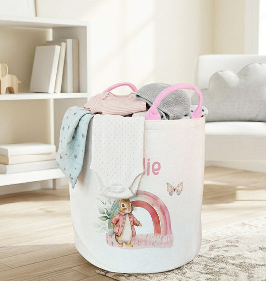 A fabric laundry hamper with pink handles sits on a rug in a bright, modern nursery. The white hamper features a colorful illustration of a penguin under a rainbow and the name 'Belle' printed on the front, with baby clothes draped over the top.