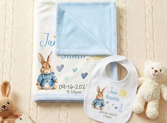 Personalized baby blanket and bib set featuring Peter Rabbit illustration and the name 'Jan Smith' with birth date and time, styled with a teddy bear on a cream cable-knit blanket.