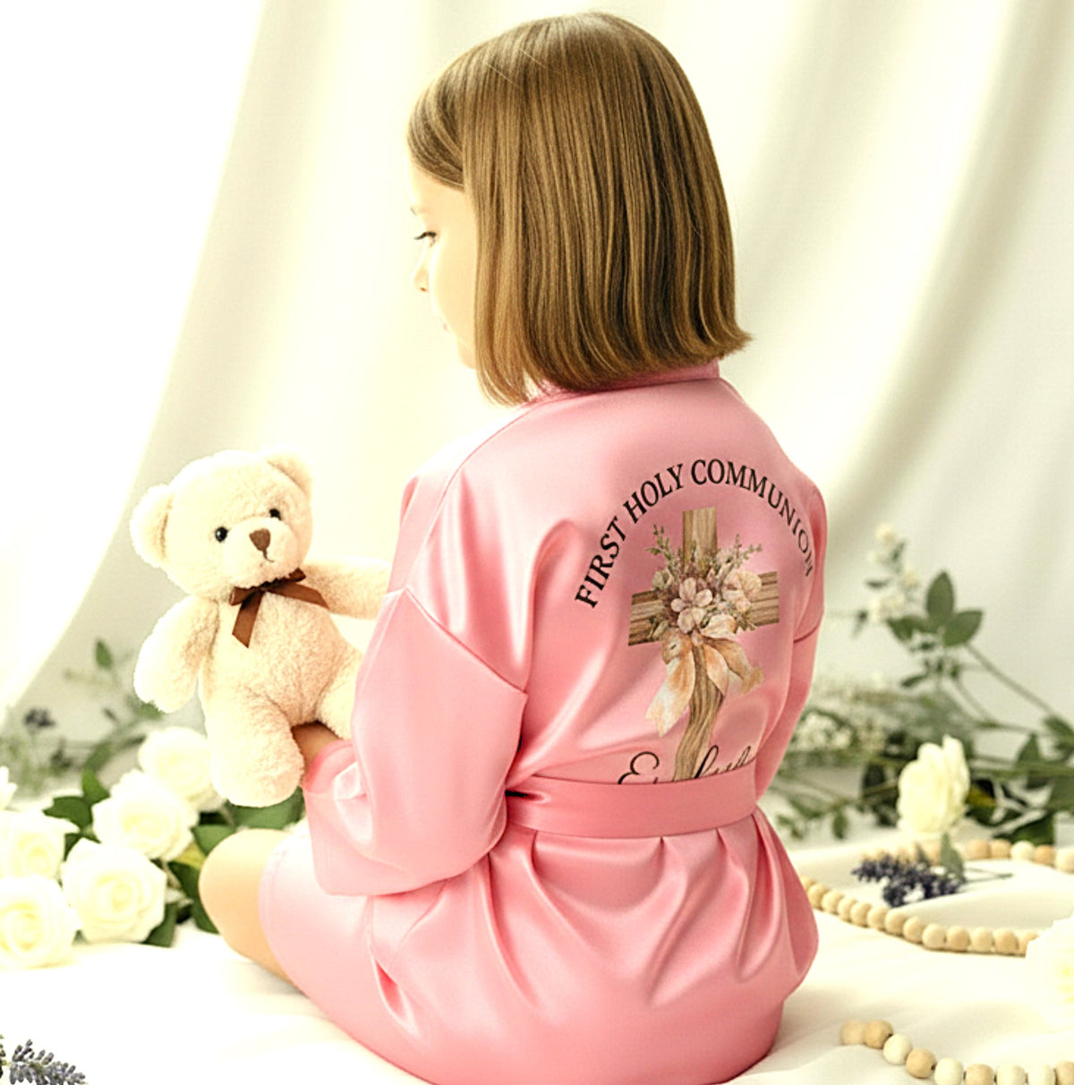 A woman seated on a white draped surface, shown from the back wearing a pink satin robe with 'FIRST HOLY COMMUNION' and a floral cross graphic on the back. She is holding a teddy bear, with white roses, beads, and greenery around her in a softly lit setting.