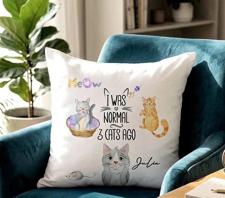 A personalized white throw pillow with cute cat illustrations and the text 'I WAS NORMAL 3 CATS AGO' with the name 'Julia', displayed on a teal velvet armchair in a cozy living room setting.