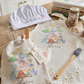 Personalized kitchen textiles displayed on a wooden table in a bright kitchen setting, including a white chef's hat labeled 'Star Baker,' a beige drawstring bag and matching bib printed with colorful dinosaur illustrations and the name 'Thomas.'.
