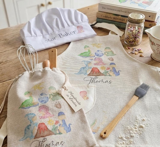Personalized kitchen textiles displayed on a wooden table in a bright kitchen setting, including a white chef's hat labeled 'Star Baker,' a beige drawstring bag and matching bib printed with colorful dinosaur illustrations and the name 'Thomas.'.